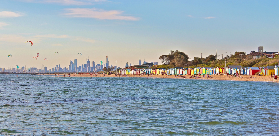 Photograph taken in the evening light from the water of Port Philip Bay, Melbourne, looking towards a row of brightly coloured bathing boxes, the city skyline in the distance and kite surfers in the distance against a blue sky with a few streaked clouds.