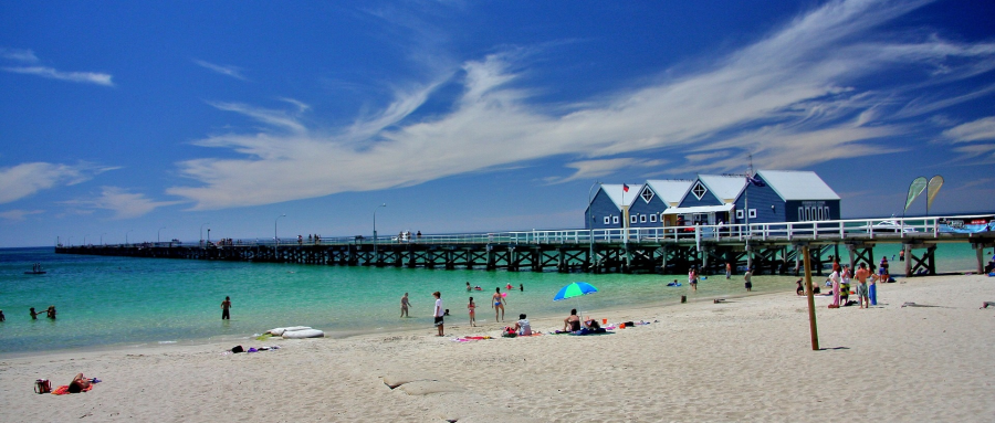 Photograph of the beach and the long jetty at Busselton, WA. The water is a clear aqua colour and there are people on the beach and in the water.